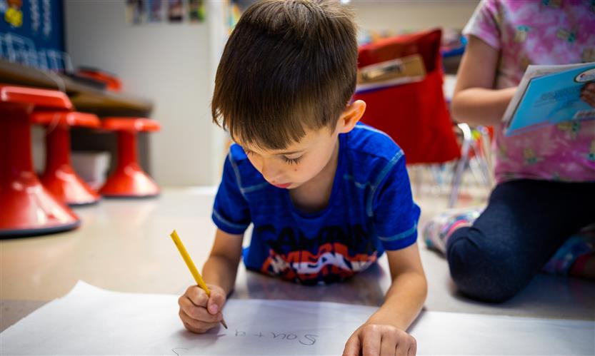 A boy wearing a blue shirt concentrates as he draws on a piece of paper